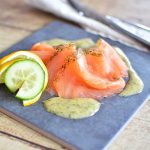 Skivad gravad lax med dill och sås på en grå tallrik, dekorerad med citronskivor och gurka.