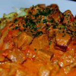 Indisk fläskgryta med bitar av fläskkött i en röd sås, serverad med grönsaker.