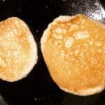 Två gyllenbruna pannkakor som steks i en svart stekpanna.