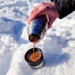 En hand häller varm choklad från en termos i en kopp som står på snö.