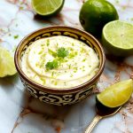 Skål med limeaioli garnerad med limezest och koriander, omgiven av limeklyftor.