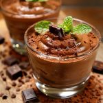 Två glas med chokladmousse toppade med mynta och chokladbitar, placerade på en träyta.