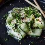 Krämig gurksallad med skivor av gurka, örter och sesamfrön på en svart tallrik.