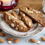 Tre skivor pepparkaksbiscotti med mandlar på en tallrik, omgivna av hela mandlar.