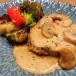 En portion grillad kyckling med svampsås, serverad med broccoli och paprika på en blå tallrik.