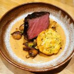En grillad köttbit serverad med grönsaker och en gul brödkaka i en djup tallrik.