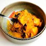 En skål med en grönsaksrätt och kött, serverad med en gaffel. Rätten har en orange sås och är garnerad med örter.